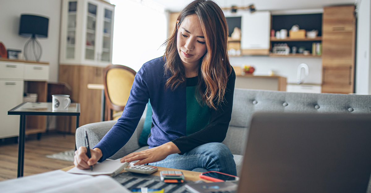 college student sitting on a couch and writing on a notebook with an open laptop computer