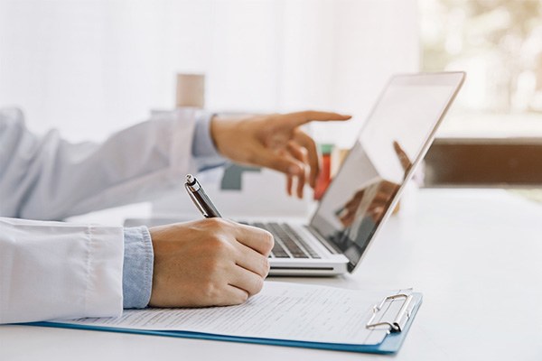 An offscreen doctor points to a laptop screen while writing on a clipboard.