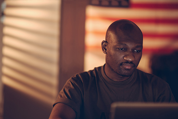 military service member looking at a computer screen and sitting in front of an American flag