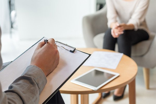 Two people sit across from each other at a coffee table while one takes notes on a clipboard.