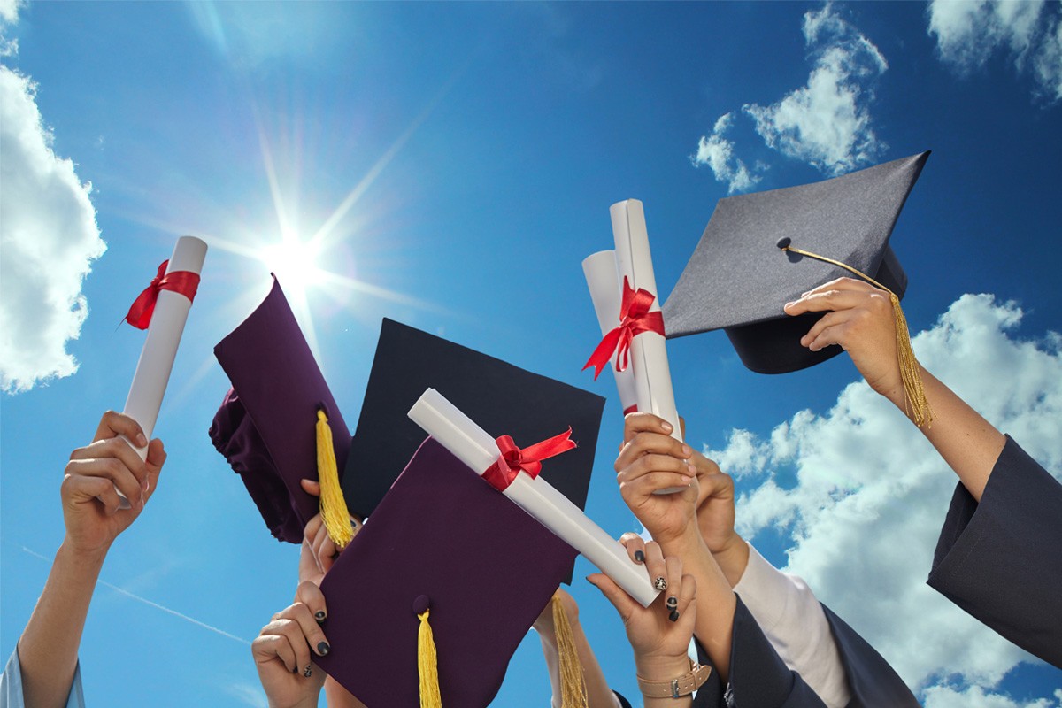 A group of hands holding diplomas and graduation caps against a cloudy sky