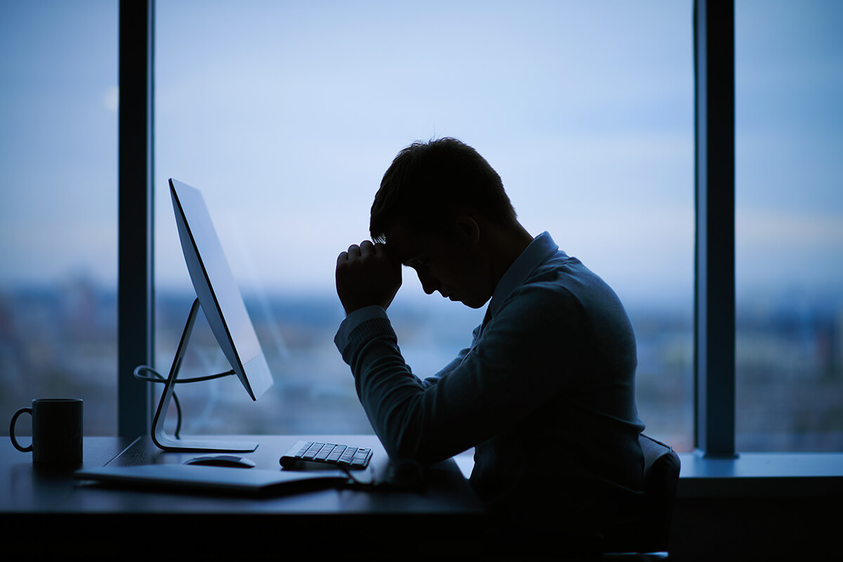 Silhouette of a worker with their elbows on their desk and their brow resting on their clasped hands.