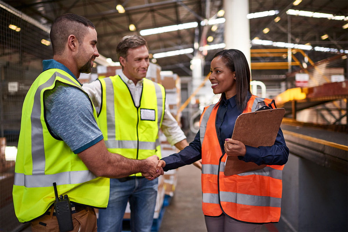 Two occupational safety and health professionals in safety vests shake hands during a meeting with a third associate.