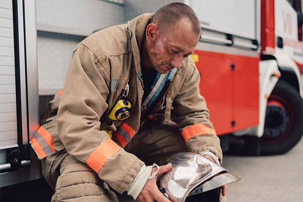 A firefighter sits on the side of a fire truck looking down at the helmet in his hands.