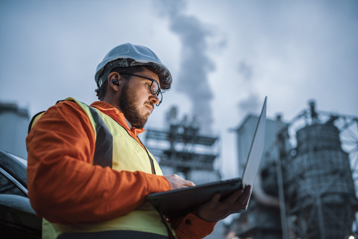 environmental manager wearing a vest and safety helmet, holding a laptop, and standing in front of a factory