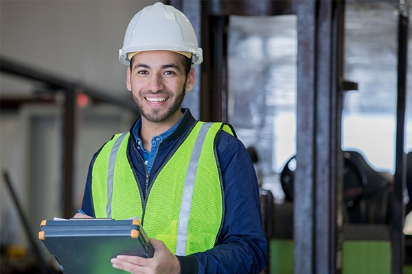 A young man in a safety vest and hard hat smiles as he holds a clip board.