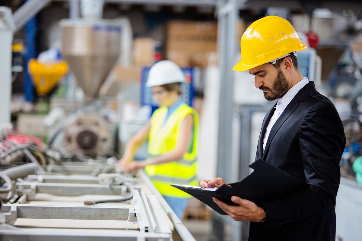 A man in a business suit and hardhat reviews a folder of paperwork on a work site.