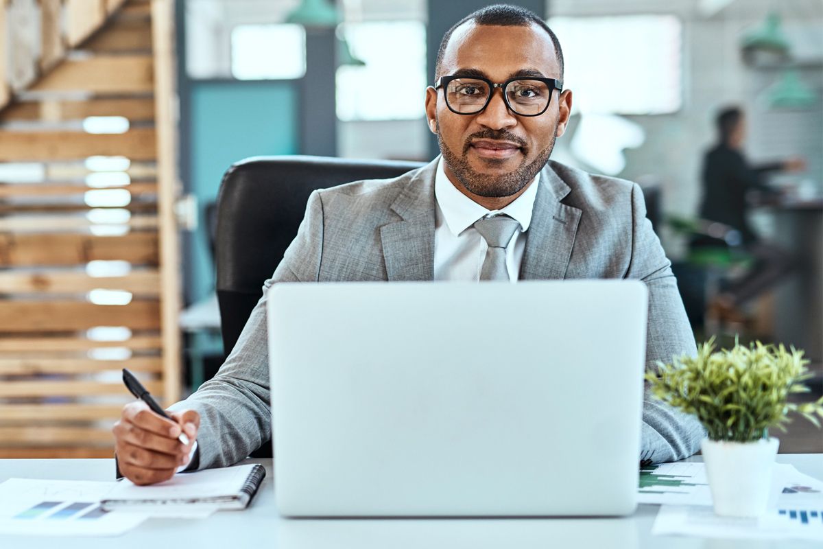 A man in a business suit sits at a desk with a notebook and laptop.