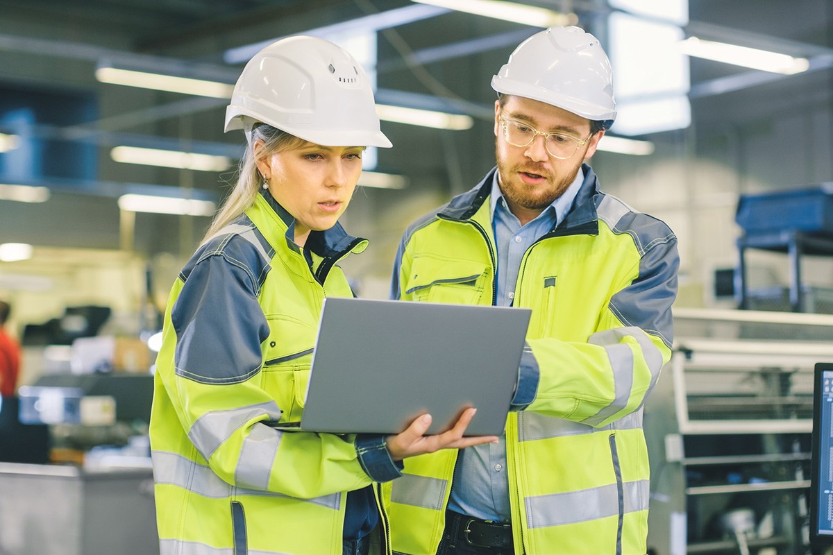 A man and woman in safety gear look at a laptop together.