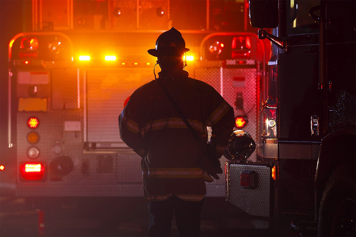 Silhouette of a firefighter standing in front of a firetruck's tail lights at night.