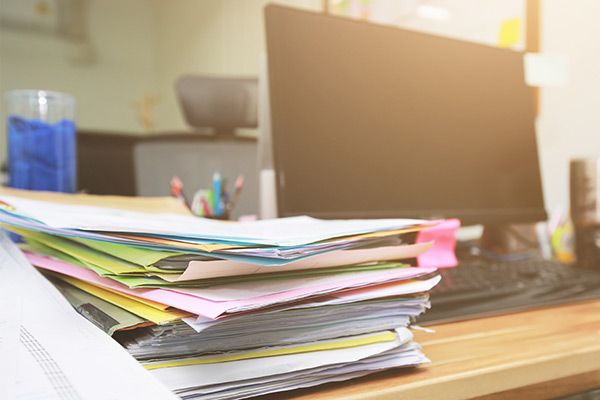 A large stack of folders and paperwork sits on a computer desk