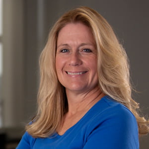 A middle aged Caucasian woman with long blonde hair smiles into the camera. She is wearing a blue shirt.