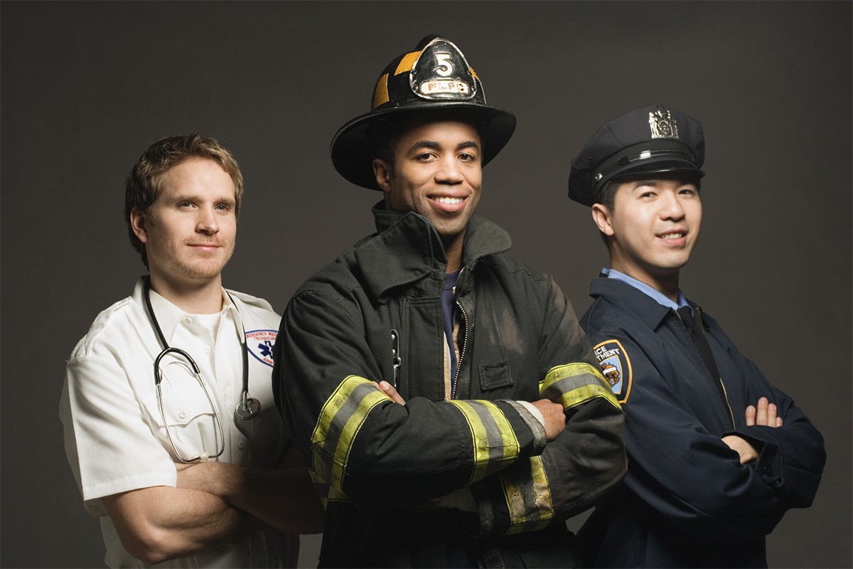 An EMT, firefighter and police officer stand together smiling with arms crossed.