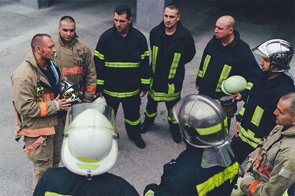 A group of firefighters stand in a circle as one speaks to them all. 