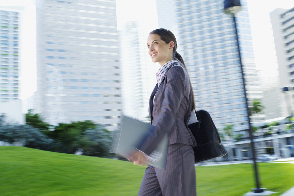 A woman in a pin striped business suit walks through a commercial park with a briefcase and files.