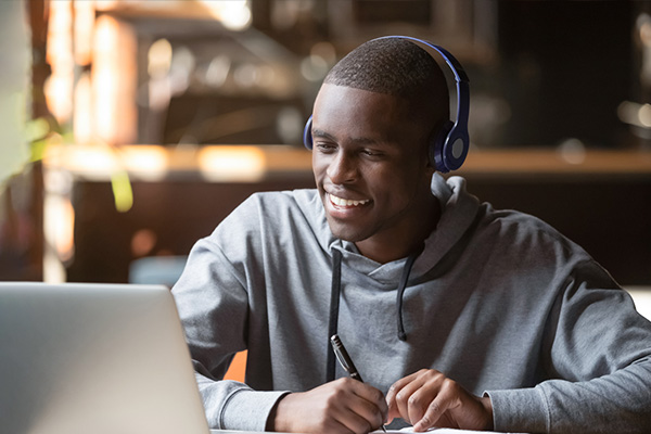 College student smiling and wearing headphones and holding a pen while looking at a laptop computer