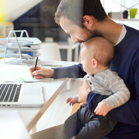 A father sits with his baby in his lap while copying information from his laptop onto a notepad.