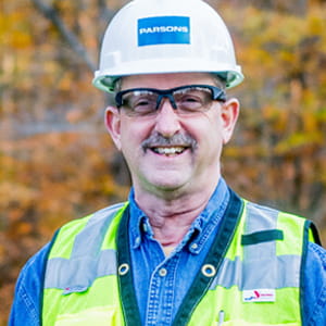 A middle aged Caucasian man smiles while wearing protective safety gear: a white helmet, glasses, and yellow vest.