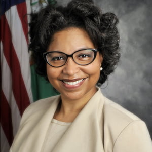 Shanna Travic is wearing glasses and a tan suit in front of an American flag.