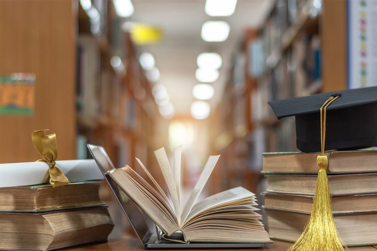 A crowded library desk with an open book resting on a laptop framed on either side by a diploma and a graduation cap