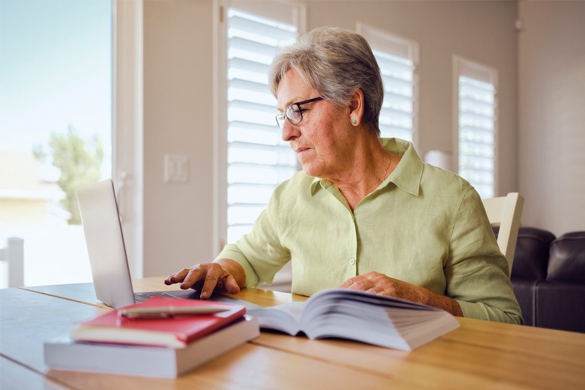 An older woman studies on a laptop with a book open at her kitchen table.