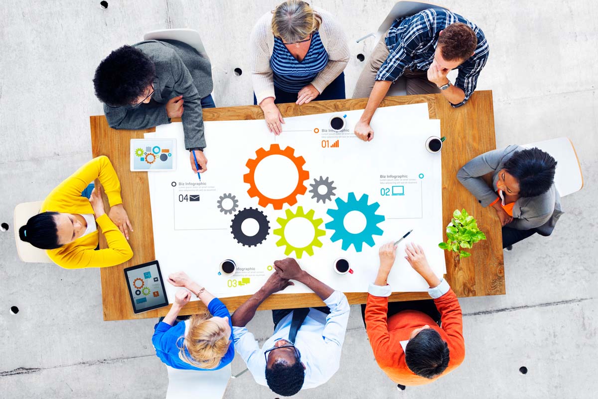 Pictured from above, eight business professionals sit at a desk and examine designs on paper and tablets