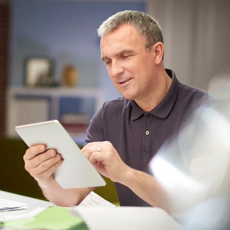 A clean shaven, middle-aged student in a dark grey polo shirt happily uses a tablet computer at his desk.