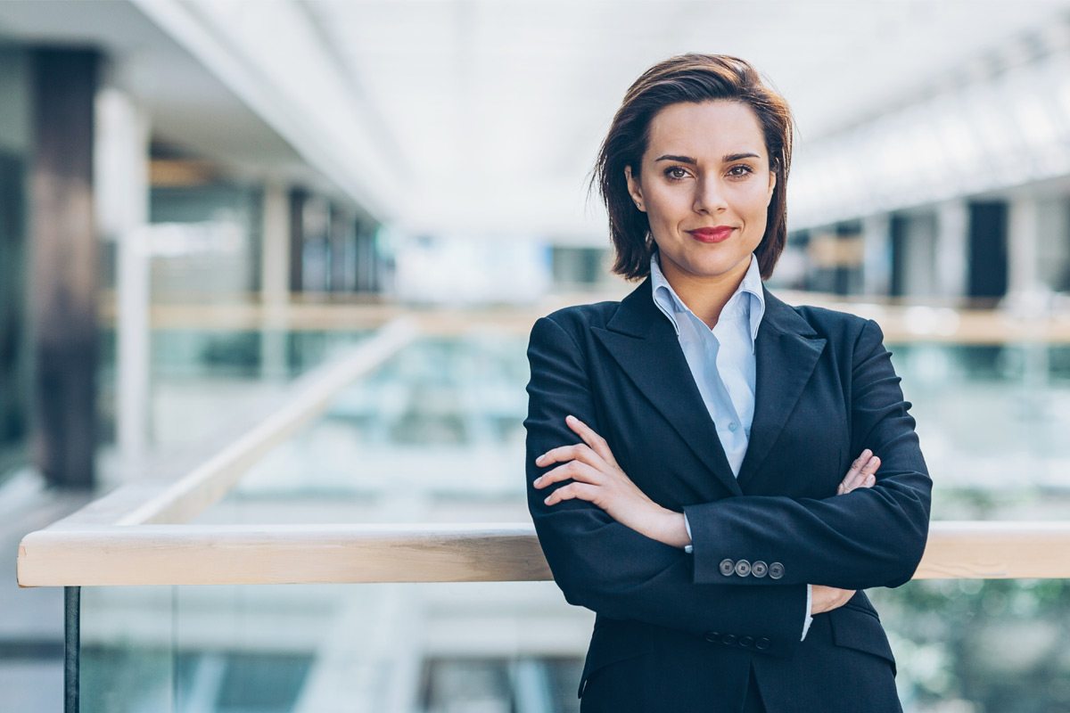 A woman in a business suit smiles confidently as she leans against a railing, arms crossed.