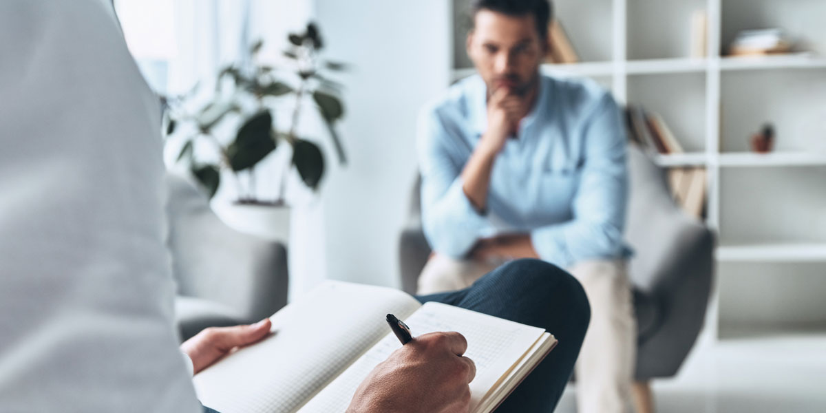 over-the-shoulder perspective of a psychologist writing in a notebook in their lap while a person in the background sits in a chair with their chin in their hand