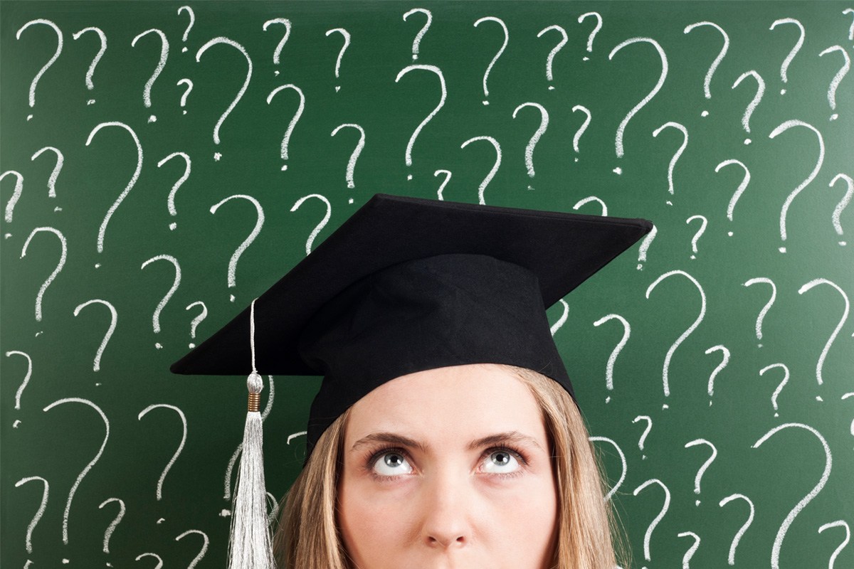 A young woman in a graduation cap stands in front of a wall covered in question marks