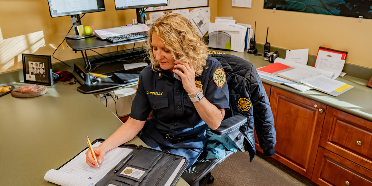 public administration professional sitting at a desk, talking on a cell phone and writing in a notebook