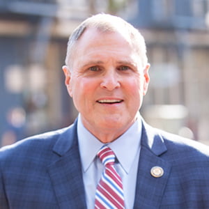 A clean shaven middle aged Caucasian man smiles slightly. He is wearing a a blue plaid suit.