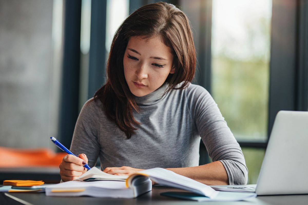 A woman takes notes while she studies with a laptop and a book.