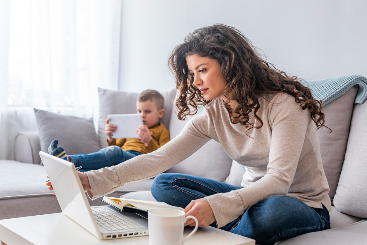 Military spouse sitting on a couch and working on a laptop while child sits next to them and plays on a tablet