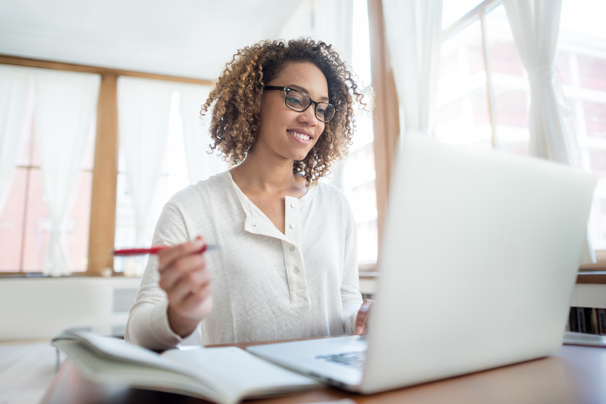 Online student smiling and sitting at desk with laptop and notebook, looking at screen, holding pen