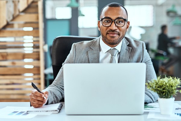 A man in a business suit sits at a desk with a notebook and laptop.