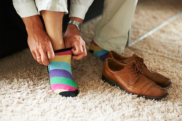 Hands pull on a brightly colored striped sock beside a pair of men's dress shoes.