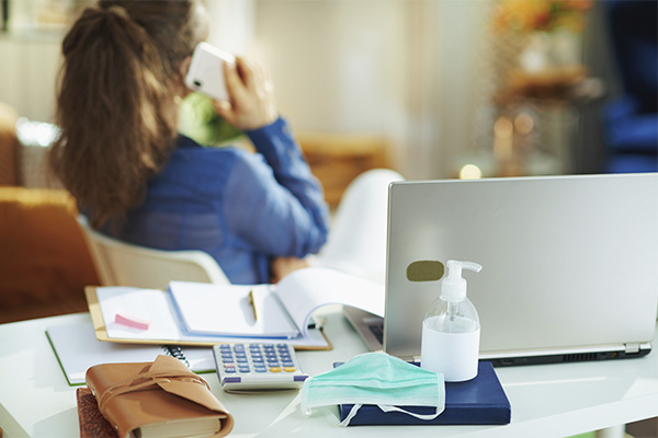 desk with office supplies, hand sanitizer and mask in foreground, person sitting and talking on phone in background