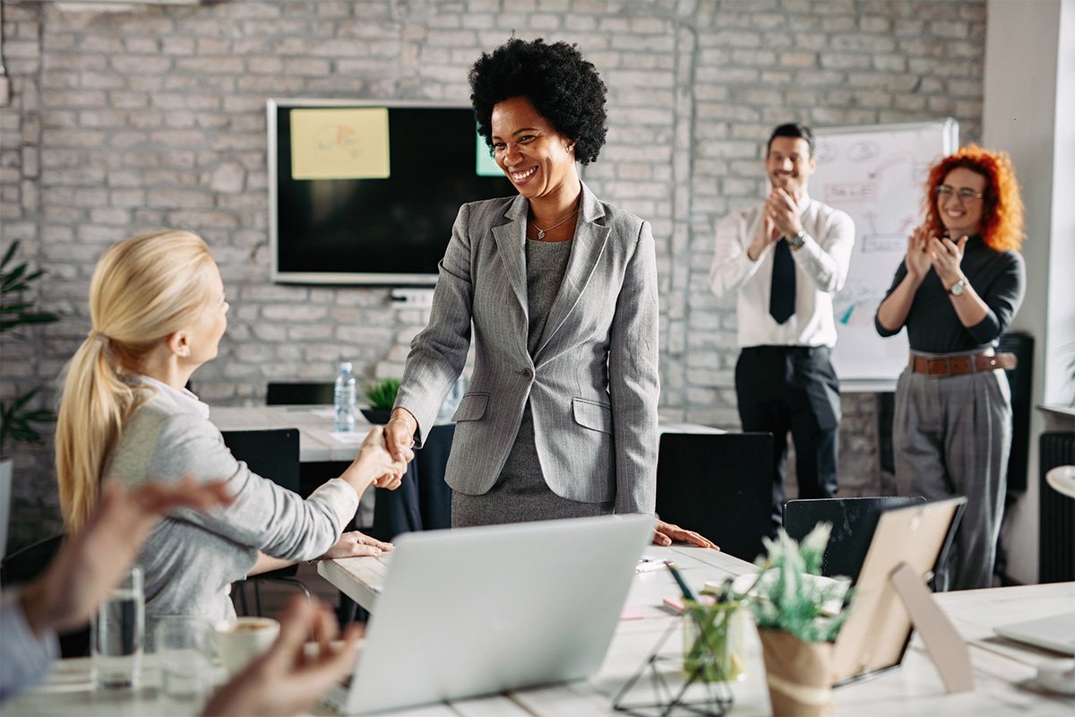 Two women in business suits shake hands while onlookers applaud.