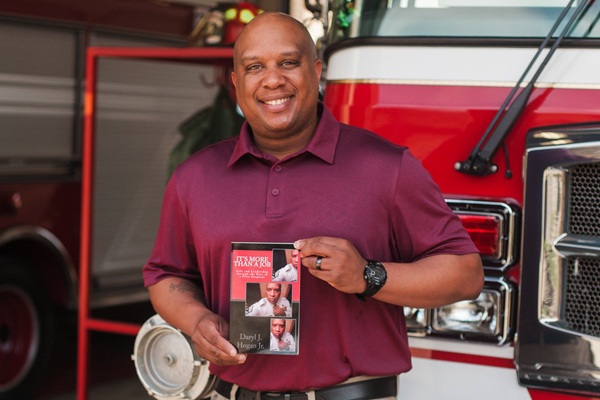 Daryl Hogan smiles in front of a fire truck as he holds his book "It's More Than a Job".