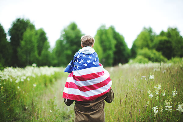 A child wrapped in an American flag like a cape rides on their parent's shoulders.