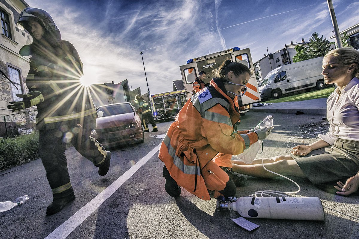 An EMT treats an injured person sitting on a street while other first responders work in the background