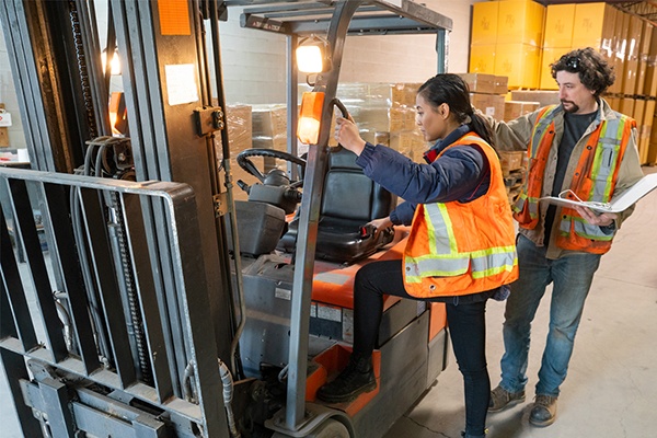 A woman in a safety vest climbs into a forklift while a man in a safety vest looks on.