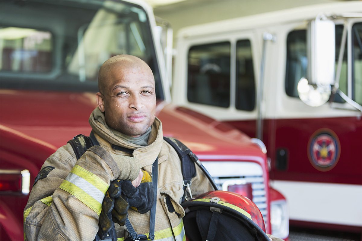 A firefighter stands in front of a fire truck holding his helmet at his side.