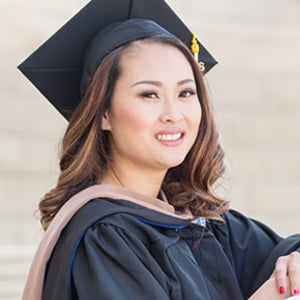 A young Asian woman with long brown hair smiles. She is wearing her cap and gown for commencement.