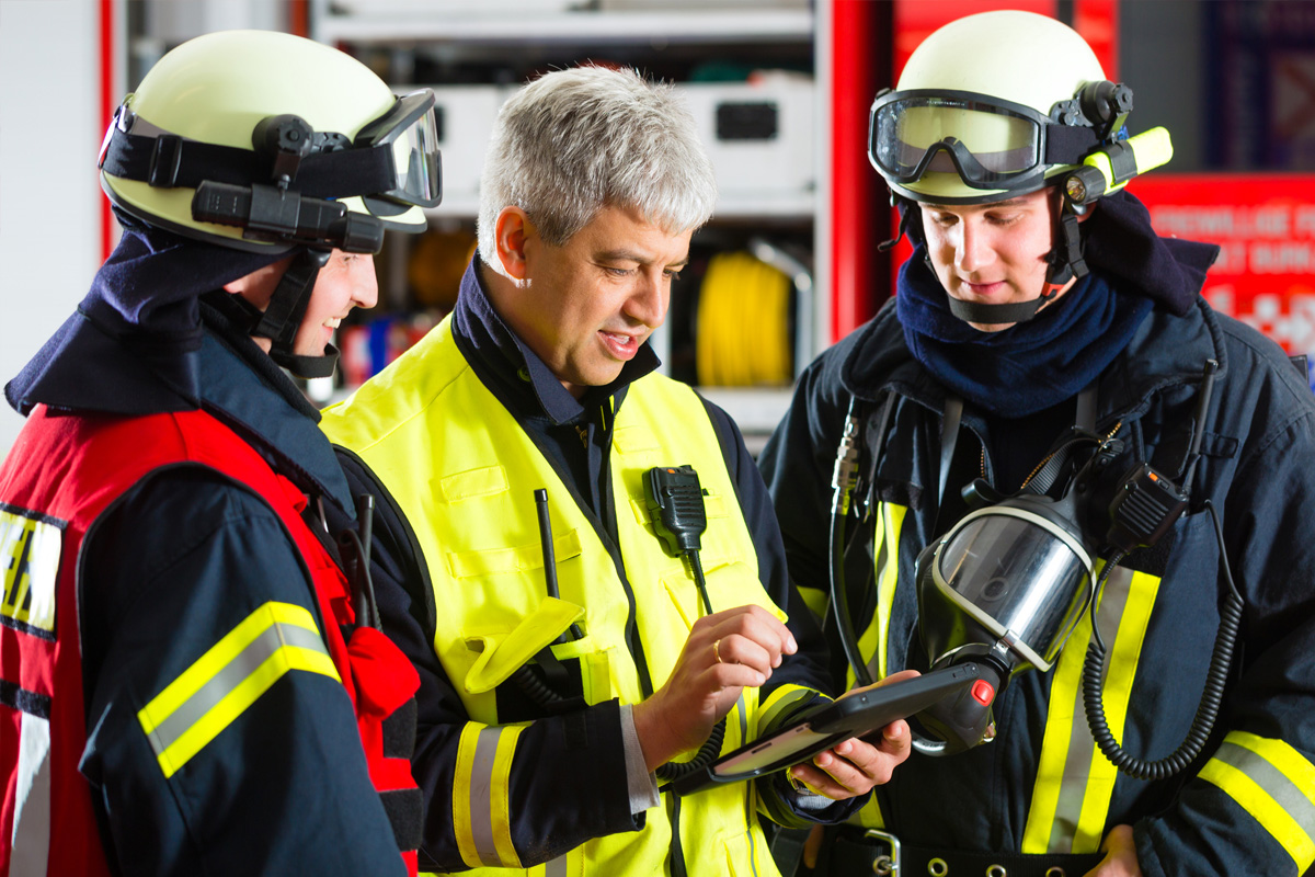 Three firefighters standing in a firehouse and smiling while reviewing information on a tablet