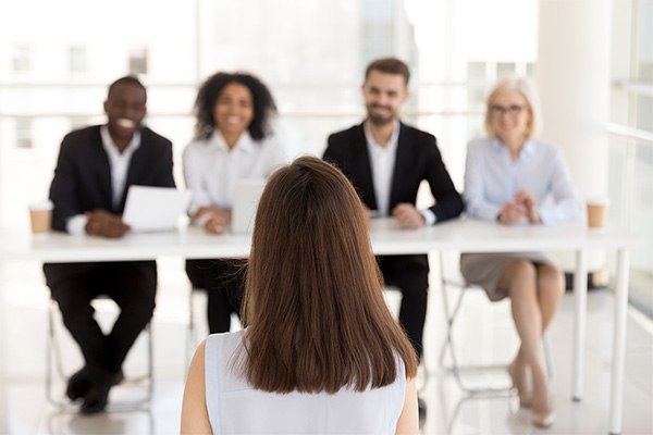 A person has their back turned to look at a panel of smiling interviewers in business dress.