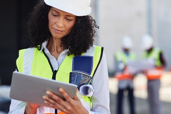 A woman in safety vest and helmet looks at a tablet.