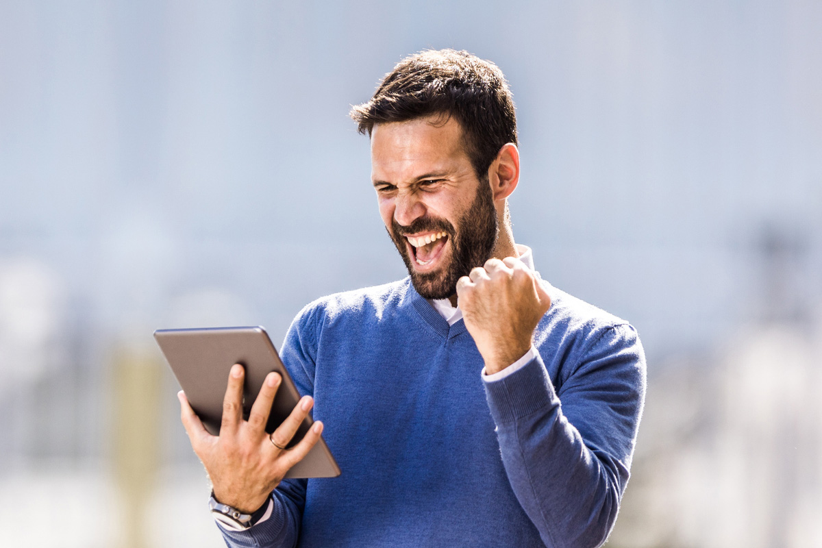 online college student standing and holding a tablet with one hand while celebrating with a fist pump in the other hand