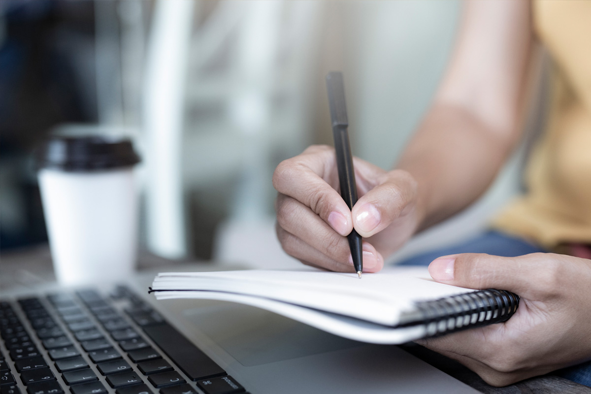 close-up image of a student's hands on a desk, one hand is holding a notebook and the other is writing in the notebook with a pen